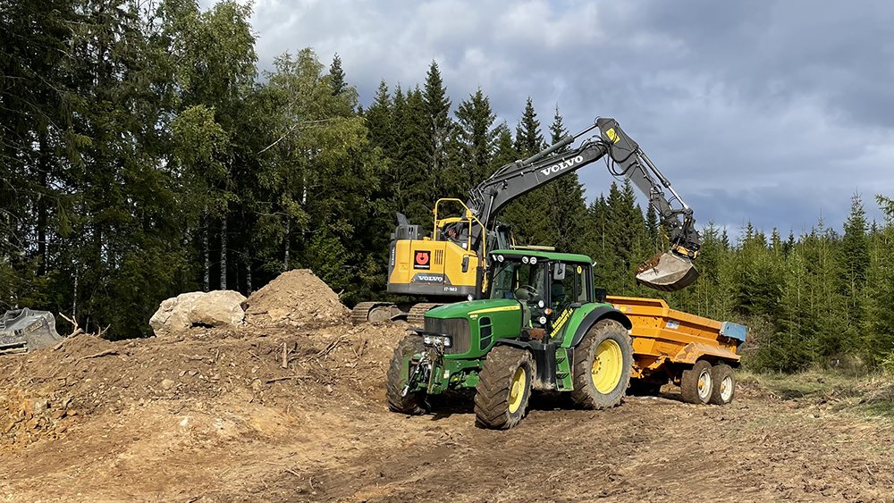 GRavemaskin og traktor med henger i arbeid på en skogsvei. Foto.